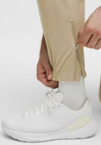 Bež sportske hlače s patentnim zatvaračima na gležnjevima, uparene s bijelim trkačkim tenisicama. Tkanina izgleda lagano i glatko, što poboljšava fleksibilnost.