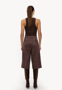 Haut marron sans manches, pantalons marron taille haute à jambes larges, texture lisse, poches arrière, portés avec des bottes marron hautes à talons.