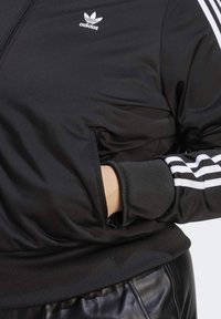 Osoba oblečená v černé bundě Adidas s bílými pruhy, ruka částečně v přední kapse, doplněná černými koženými kalhotami.