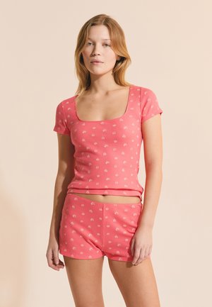 Jeune femme portant un haut rose côtelé à manches courtes et un short assorti avec un petit motif floral blanc, posant devant un fond beige.