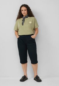 T-shirt verde oliva chiaro con maniche arrotolate e grafica solare; abbinato a pantaloni cropped scuri e una sciarpa nera a fantasia. Scarpe nere senza lacci.