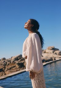 Light, flowing white blouse with wide sleeves and crocheted skirt. Model stands by water, with rocks and clear blue sky in the background.