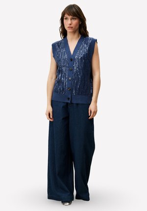 Gebreid mouwloos vest in donkerblauw met paillettenversiering, gecombineerd met wijde corduroy broek in bijpassende kleur. Knopen aan de voorkant.