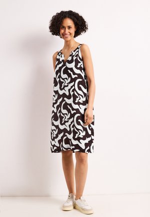 Kvinde smilende, stående, iført ærmeløs sort-hvid abstrakt mønstret kjole og hvide sneakers mod ensfarvet baggrund.