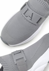 Graue Sportschuhe mit einem gestrickten Obermaterial, ausgestattet mit einem breiten grauen Schnallenriemen und einer weißen Außensohle, die eine strukturierte Oberfläche und minimale Details aufweisen.