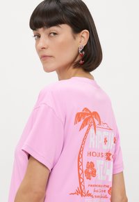 Roza majica s kratkimi rokavi, ki na hrbtni strani vsebuje velik oranžen grafični vzorec s palmami in cvetjem. Gladek material in sproščen kroj.