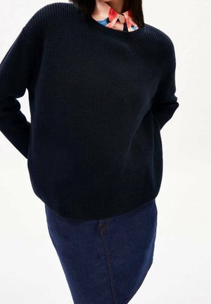 Personne portant un pull en maille noir par-dessus une chemise à col à motifs multicolores et une jupe en denim bleu foncé, les mains derrière le dos.