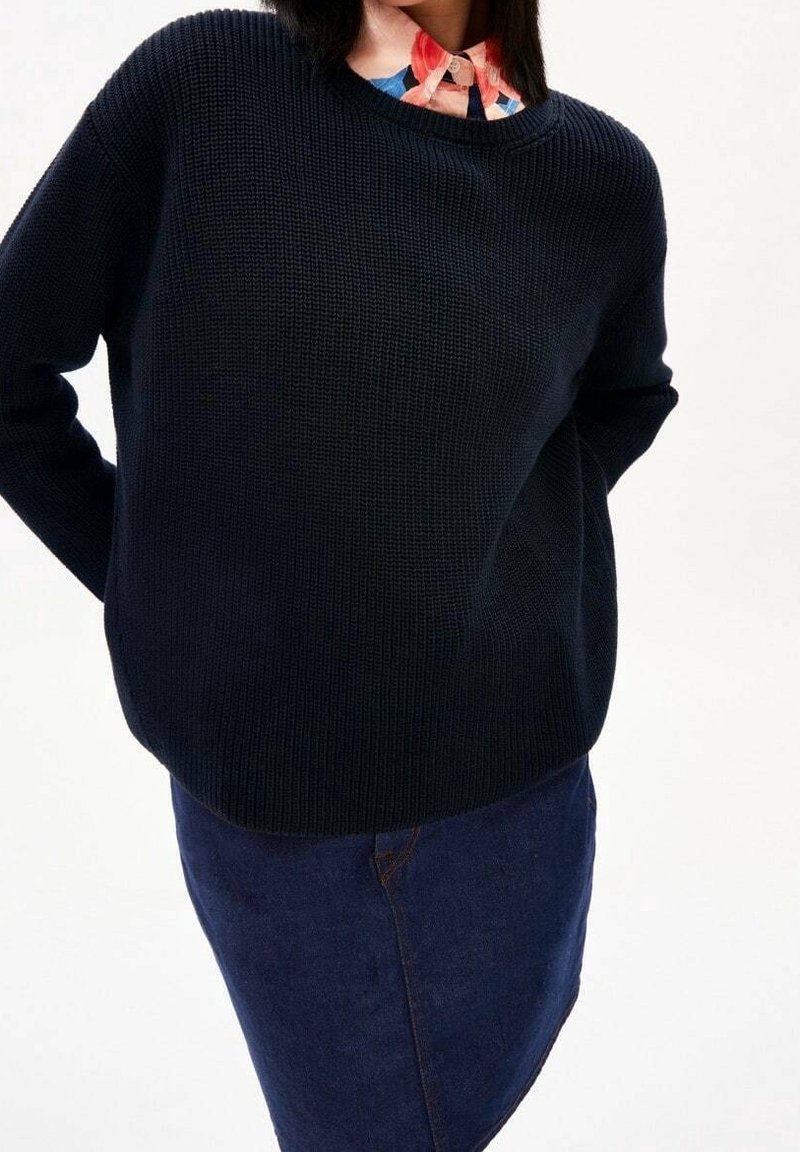 Personne portant un pull en maille noir par-dessus une chemise à col à motifs multicolores et une jupe en denim bleu foncé, les mains derrière le dos.
