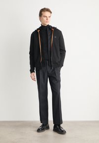 Black zip-up hoodie with orange accents, worn over a black turtleneck and trousers, paired with black shoes. Smooth fabric, relaxed fit.