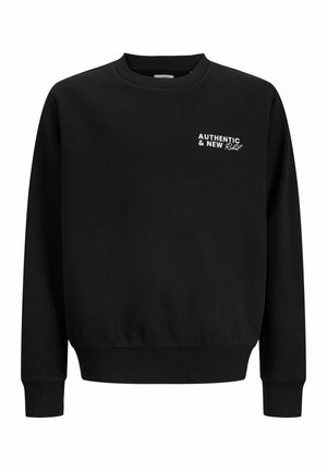 Garçon blond debout, une main dans la poche, portant un sweat-shirt noir, un pantalon beige et des baskets blanches sur un fond clair uni.