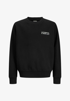 Garçon blond debout, une main dans la poche, portant un sweat-shirt noir, un pantalon beige et des baskets blanches sur un fond clair uni.