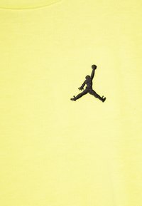 Camiseta amarilla con un logo Jumpman bordado en negro en el lado izquierdo del pecho. El material parece suave y tiene una textura lisa.