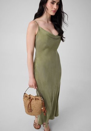 Frau in olivgrünem ärmellosem Kleid, die eine geflochtene Strohtasche hält, trägt braune Riemchensandalen und steht vor einem schlichten Hintergrund.