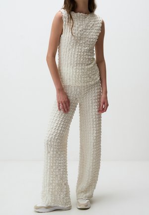 Mujer con top blanco sin mangas y texturizado y pantalones anchos a juego con zapatillas beige y blancas sobre un fondo liso.
