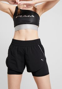 Camiseta deportiva corta negra con banda plateada que presenta el logo "PUMA"; combinada con pantalones cortos negros, detalles de malla y cintura elástica.