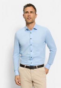 Chemise bleu clair à manches longues en tissu lisse, avec des boutons foncés et un col classique. Associée à un pantalon beige et une ceinture noire.