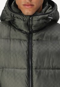 Tamnozeleni puffer sako s teksturiranim uzorkom, visokim ovratnikom, podesivom kapuljačom i crnim zatvaranjem na patentni zatvarač sprijeda.