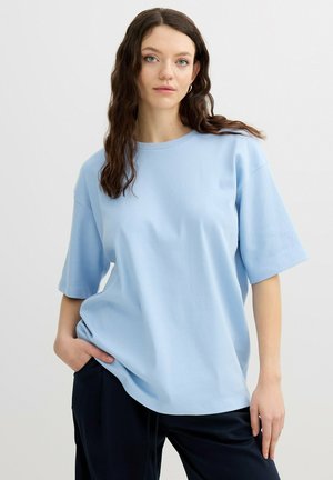 Mujer con cabello castaño ondulado lleva una camiseta azul claro de manga corta con cuello redondo, pantalones negros, de pie con una mano en el bolsillo contra un fondo liso.