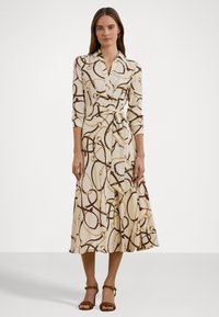 Long-sleeve, collared dress in cream fabric featuring a brown and gold chain pattern, with a tie waist and flowing skirt. Brown strappy sandals.