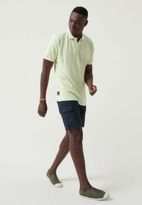 Camisa polo verde claro, de mangas cortas, combinada con pantalones cortos cargo azul marino. El modelo lleva zapatillas de deporte verdes oliva, posando contra un fondo neutro.