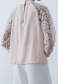 Blouse en tissu rose clair avec manches longues en dentelle texturée, fermeture au dos en forme de goutte, et corps lisse. Finition soyeuse avec un drapé doux.