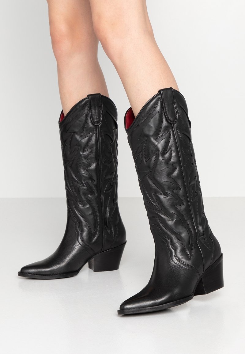 Jambes portant des bottes de cow-boy en cuir noir montantes jusqu'aux genoux, avec des bouts pointus, des talons empilés et des détails embossés de style occidental.