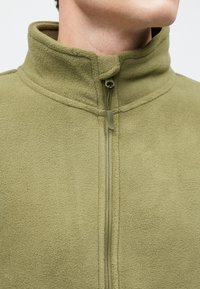 Olivovo zelená fleeceová bunda so stojacím golierom, celopredným zipsom a jemnou textúrou. Má prešívanie pozdĺž zipsovej línie.