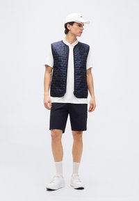 Άνδρας μοντέλο που φοράει μπλε σκούρο καπιτονέ γιλέκο πάνω από λευκό πόλο πουκάμισο, μπλε σορτς, λευκές κάλτσες, λευκά αθλητικά παπούτσια και λευκό καπέλο, στέκεται και κοιτάζει πλάγια.