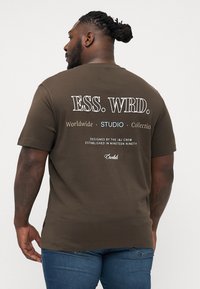 Brun bomuld t-shirt med korte ærmer, med hvid tekst på ryggen: "ESS. WRD. Worldwide STUDIO Collectio." Designet af J&J Crew.