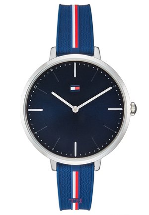 Montre ronde en argent avec cadran bleu marine, index horaires blancs minimalistes, logo Tommy Hilfiger et bracelet bleu à rayures rouges, blanches et bleues.