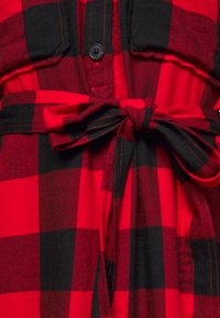 Camicia a quadri rossa e nera con bottoni frontali, due tasche sul petto e cintura da annodare in vita. Il tessuto appare morbido e texture.
