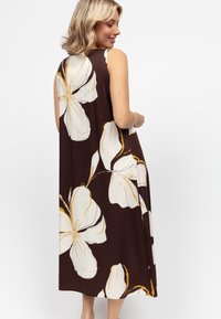 Sukienka maxi bez rękawów w ciemnobrązowym materiale z dużymi białymi kwiatami z żółtymi akcentami. Boczne rozcięcia poprawiają przepływ powietrza i umożliwiają swobodne poruszanie się.
