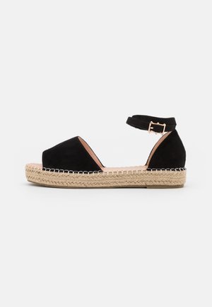Sandalia tipo espadrille de ante negro con tiras en el tobillo y hebilla, suela plataforma envuelta en yute, punta abierta y talón cerrado sobre fondo blanco.