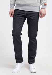 Hombre con jeans oscuros de corte slim, sudadera gris y zapatillas blancas, de pie con las manos relajadas a los lados sobre un fondo blanco.