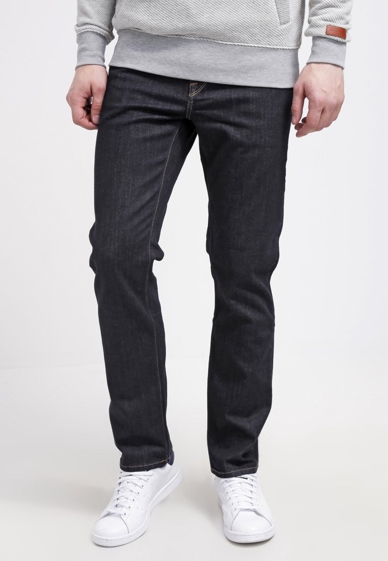 Hombre con jeans oscuros de corte slim, sudadera gris y zapatillas blancas, de pie con las manos relajadas a los lados sobre un fondo blanco.