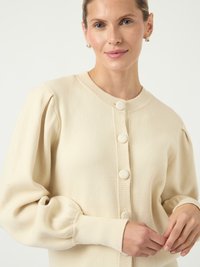 Kreminės spalvos kardiganas su pūkuotomis rankovėmis, raukiniais ant riešo ir penkiomis apvaliomis sagomis priekyje. Minkšta, tekstūruota mezgimo medžiaga.