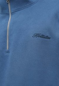 Modrá fleecová bunda na zips s vysokým golierom, s textúrovanou tkaninou a vyšitým logom "Hollister" v tmavomodrej farbe. Strieborný zips.