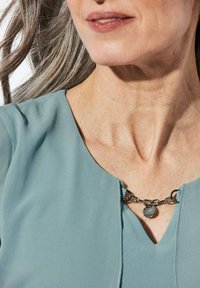 Lichtblauwe blouse met een V-hals, voorzien van een metalen kettingaccent met een ronde hanger aan de hals, gladde textuur en zachte stof.