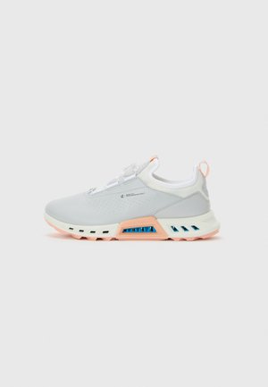 Sapatilha atlética cinza claro com detalhes em pêssego e azul na sola, design slip-on e sistema de atilhos com botão, exibida em perfil lateral contra um fundo branco.