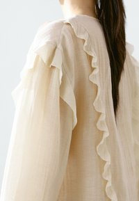 Beige blouse with ruffled sleeves made from lightweight fabric. Soft texture, loose fit, and subtle sheer quality.