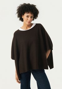 Bruine oversized top met een witte kraag, gemaakt van zachte stof. Kenmerken zijn brede mouwen en zijopeningen voor meer bewegingsvrijheid. Draagt men met donkere jeans.