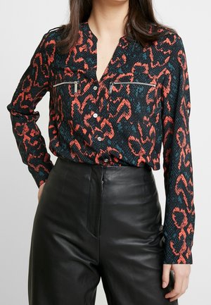 Mujer con camisa de botones estampada en negro y naranja, metida dentro de pantalones de cuero negros de talle alto, de pie con una mano en la cadera.