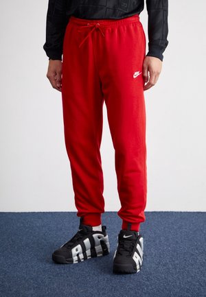 Homme portant un pantalon de survêtement rouge Nike avec un logo blanc et des baskets Nike Air noires, debout sur un tapis bleu contre un mur blanc.