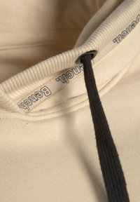 Sudadera con capucha beige con superficie texturizada, con cordón negro y etiqueta impresa en el interior del cuello. Sutiles detalles acanalados.