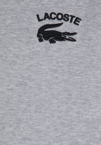 Šedé bavlněné tričko s vyšívaným černým logem krokodýla a nápisem "LACOSTE" nahoře v černé barvě, s texturovaným povrchem.
