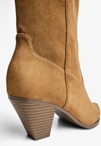 Brown suede ankle boot with a pointed toe and 3-inch conical wooden heel featuring horizontal stripes. Smooth, soft texture with stitching detail.