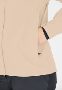 Beige vattentät jacka med en mjuk textur, fickor på sidan med dragkedja och svarta manschetter, tillsammans med mörka byxor.
