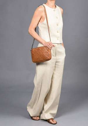 Beige ärmelloses Oberteil mit runden Knöpfen, kombiniert mit weit geschnittenen Hosen. Eine braune geflochtene Handtasche und flache Sandalen runden das Outfit ab.
