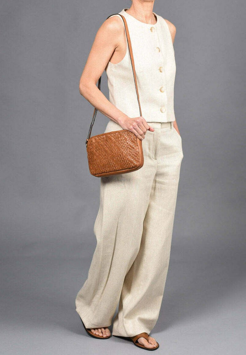 Beige mouwloos topje met ronde knopen, gecombineerd met wijde broekspijpen. Een bruine geweven handtas en platte sandalen maken de look compleet.