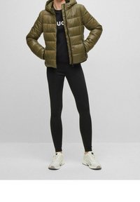 Giubbotto imbottito in nylon verde oliva con cappuccio e design a cuciture orizzontali. Indossato sopra una maglietta nera e abbinato a leggins neri e sneakers bianche.